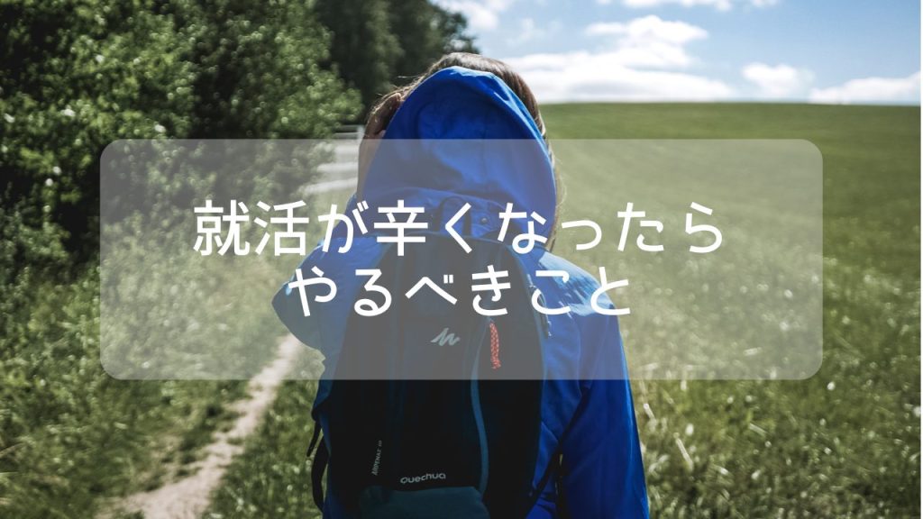 青空と広い草原の中に、青いパーカーと黒いリュックを背負ったブロンドヘアの女性がフードをかぶって向こうへ歩いている